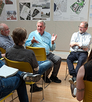 Members from local agencies and firms talking with each other. They are sitting on yellow chairs in the spacious, dome-lit, white-board adorned Jury Crit room in Marshall Hall.