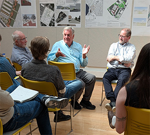 Members from local agencies and firms talking with each other. They are sitting on yellow chairs in the spacious, dome-lit, white-board adorned Jury Crit room in Marshall Hall.