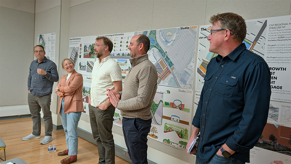 Five leaders from the Central New York community stand next to each other and address students. They are flanked by informative wall posters that explain landscape design and possibilities