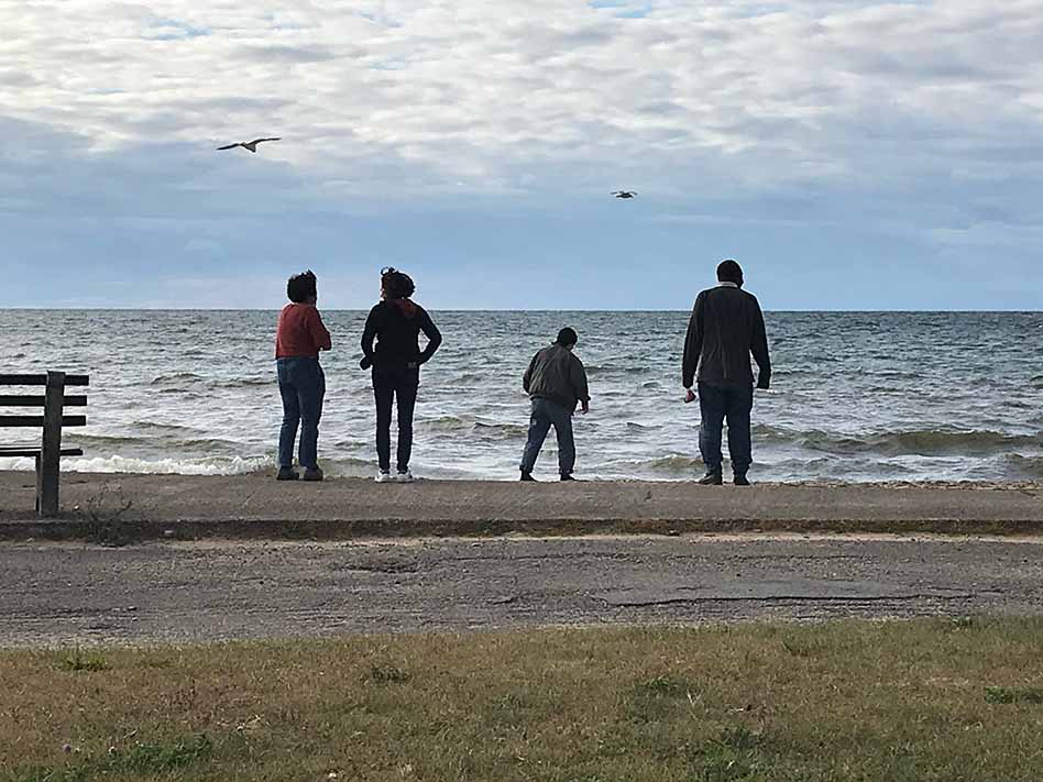 Four students and faculty at the beach with two seagulls flying overhead. The water is white capped and the students are facing the water, with their backs to the camera.