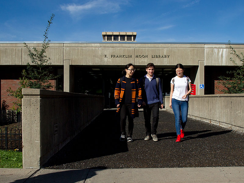 Three students walking towards the camera outside of the Moon Library entrance.