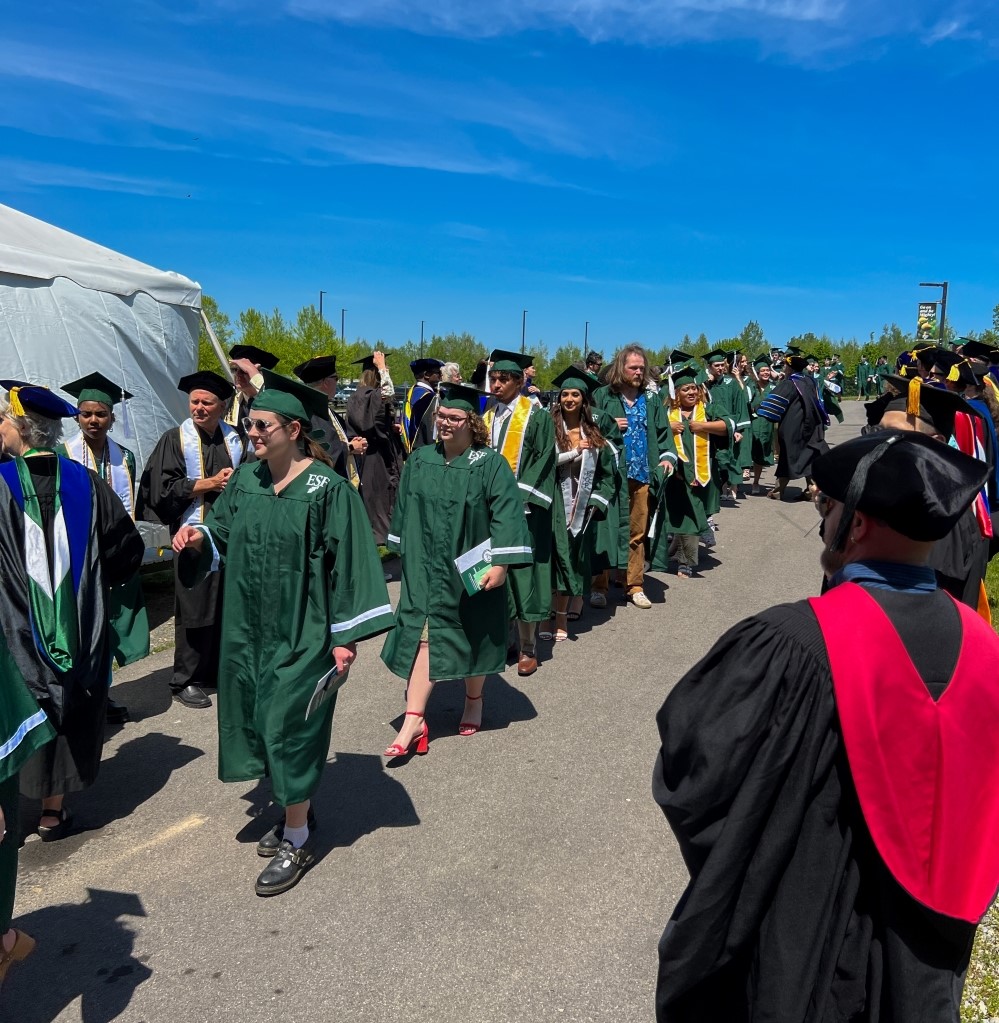 Students in green robes and graduation caps surrounded on both sides of the path by older adults with black graduation caps.