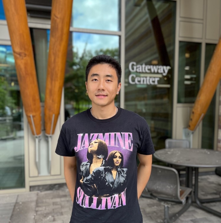 Man standing in front of a building with large wooden beams. The building has a sign that says Gateway Center. The man is wearing a Jazmine Sullivan t-shirt.