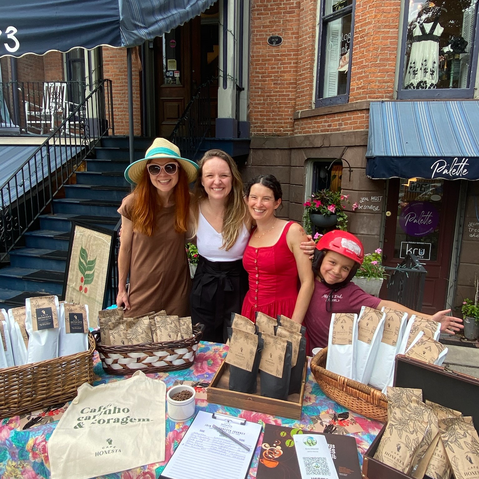 Four woman and kid standing in front of a table filled with coffee bags and merch. Behind them in a brick and metal building circa 1800s architecture.