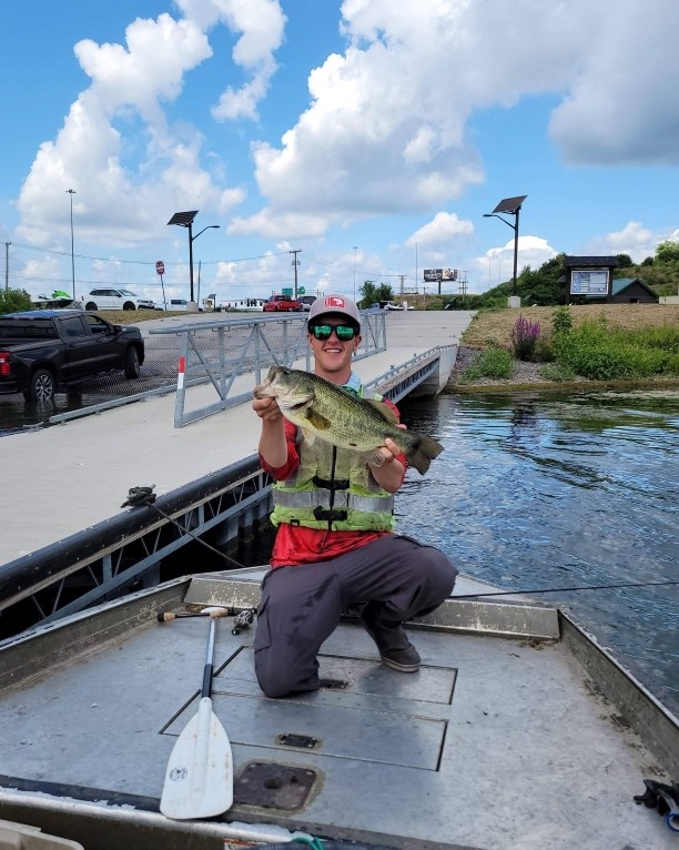Man on a boat with a green life jacket, sunglasses and a white hat holding a fish.