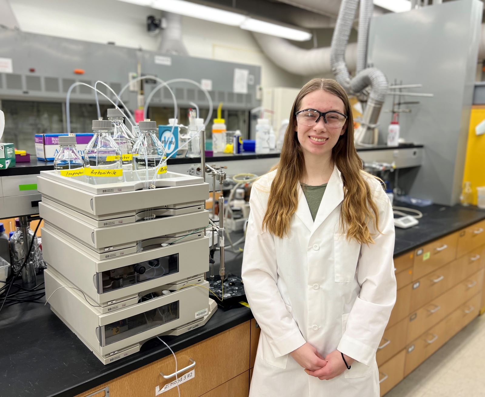Woman with safety glasses and a lab coat on standing next to a machine that has several beakers with tubes coming out of them.