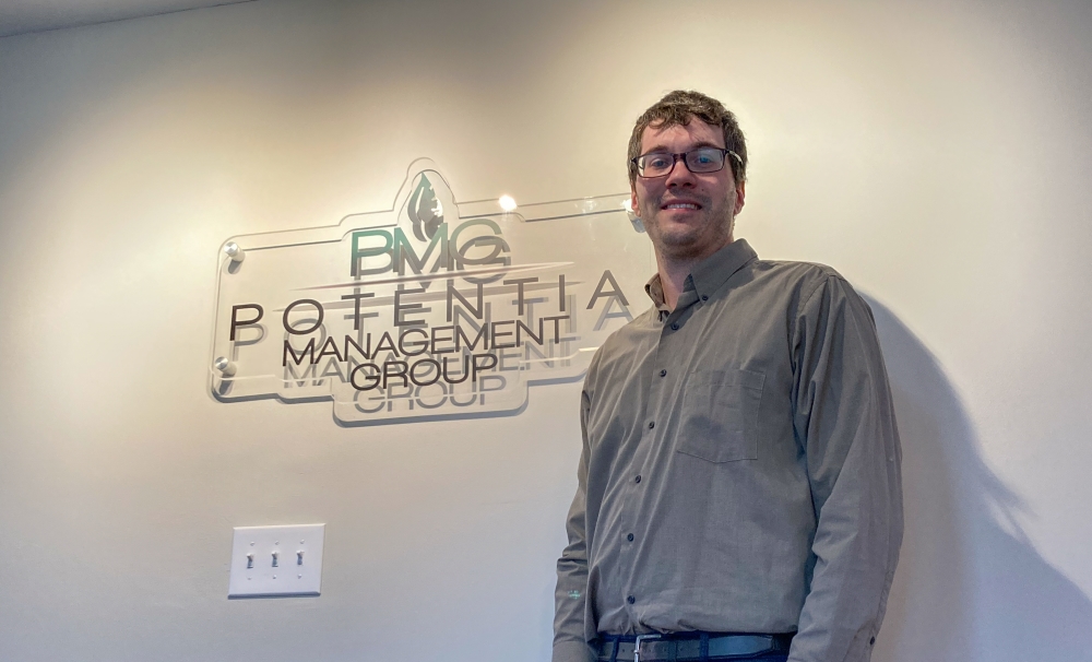 Man in a grey button down shirt standing in front of a sign that says PMG Potentia Management Group.