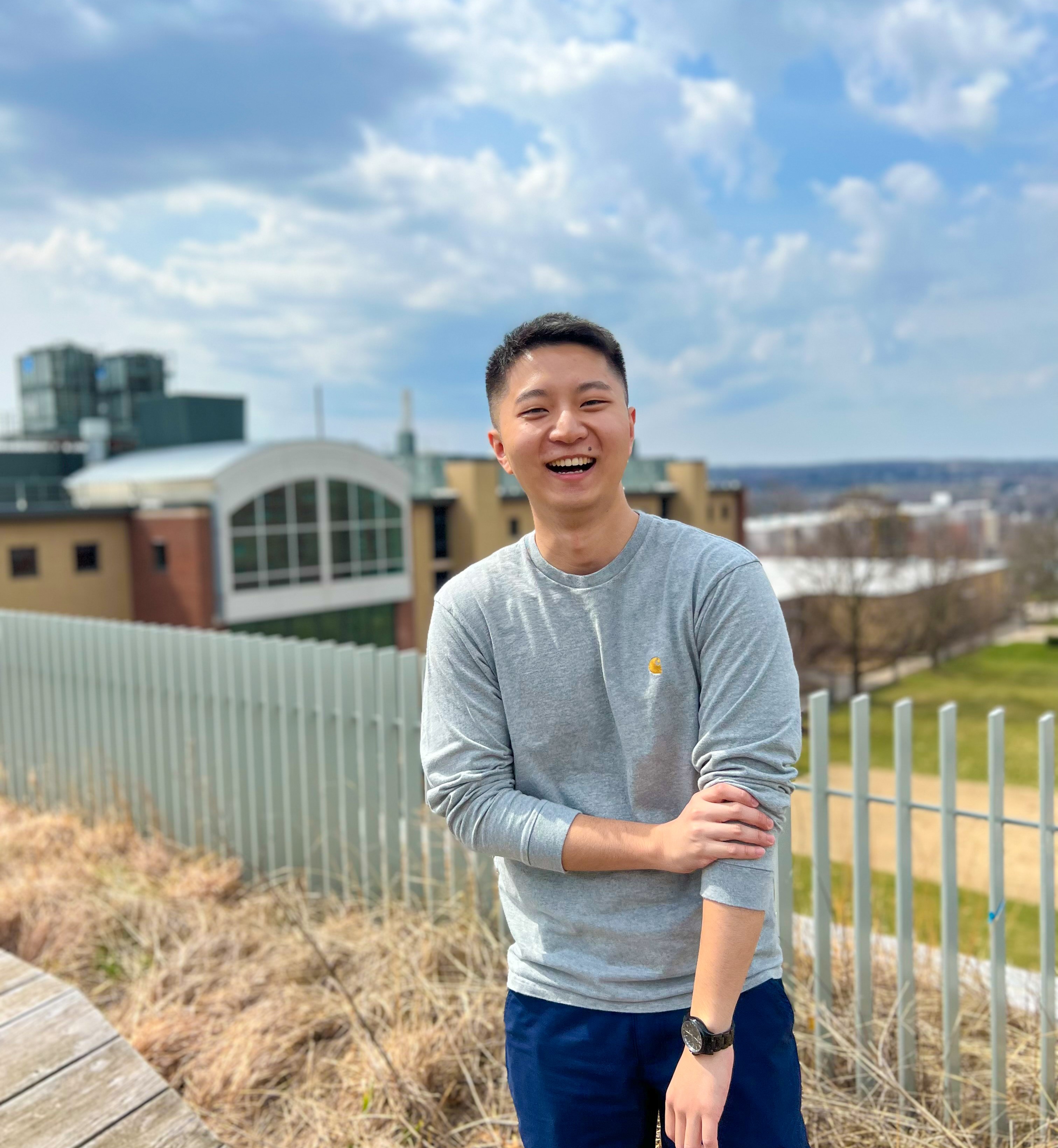 Man with a grey shirt standing outside with some buildings, a green field, and blue skies behind him. There is a fence that separates the horizon from the platform.