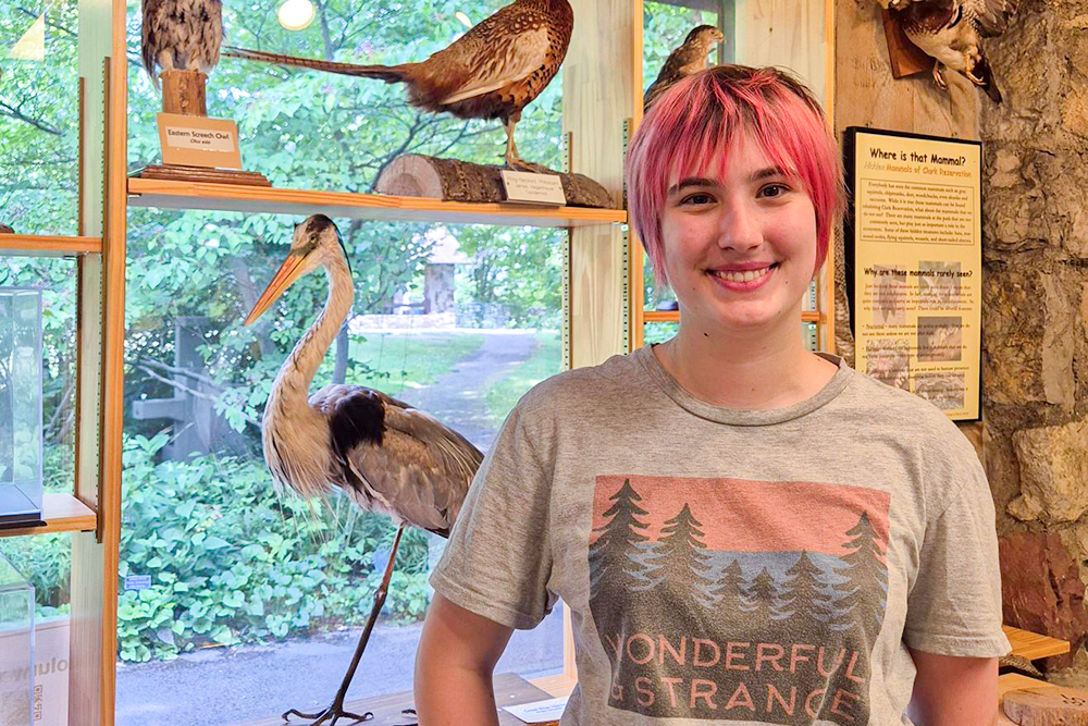 Young woman with short pink hair and T-shirt with trees standing in front of bird display.