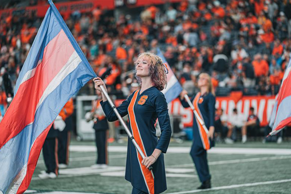 Young woman with shoulder length hair wearing blue and orange band uniform holding a colorguard flag that is also blue and orange.