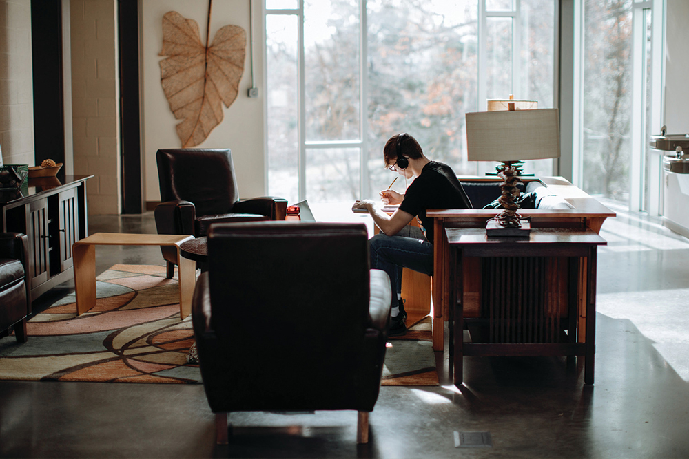 Student sitting on a wood-rimmed couch in a lounge featuring two chairs, big windows and an area rug.