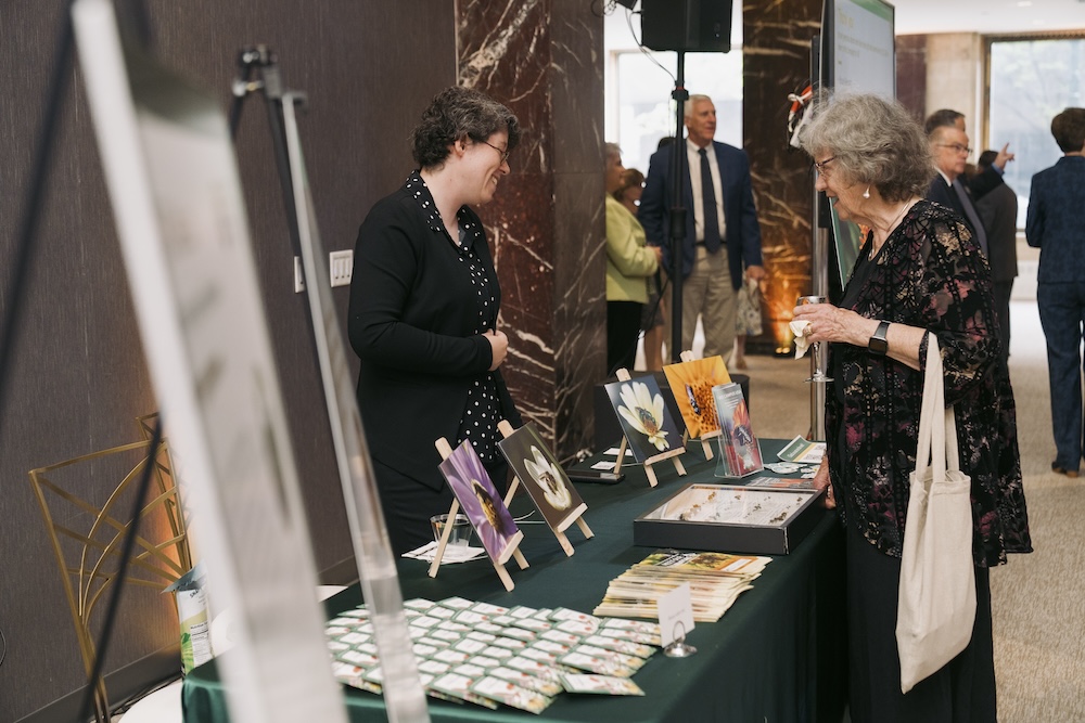 Woman wearing black talks with woman holding tote bag over a table display.