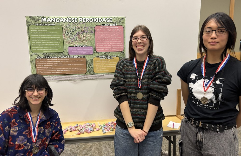 Three women in front of a poster. One woman is sitting, all are wearing medals for winning best enzyme.