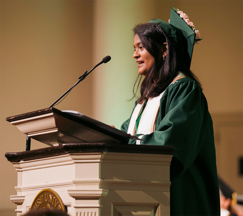 Abhinaya Polaka addresses a commencement in graduation regalia.