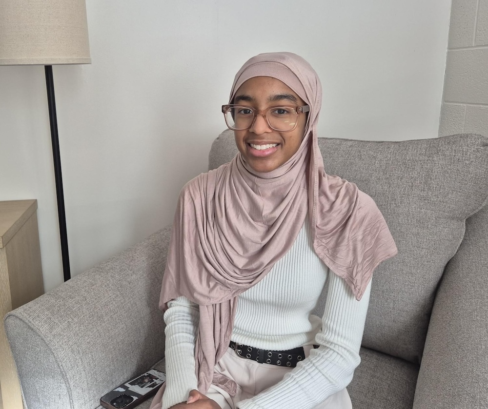 A person wearing a light pink hijab and glasses sits on a gray couch. They are dressed in a white ribbed sweater and light-colored pants, with a black belt around the waist. A wooden side table with a lamp is visible to the left, and the background features a plain white wall.