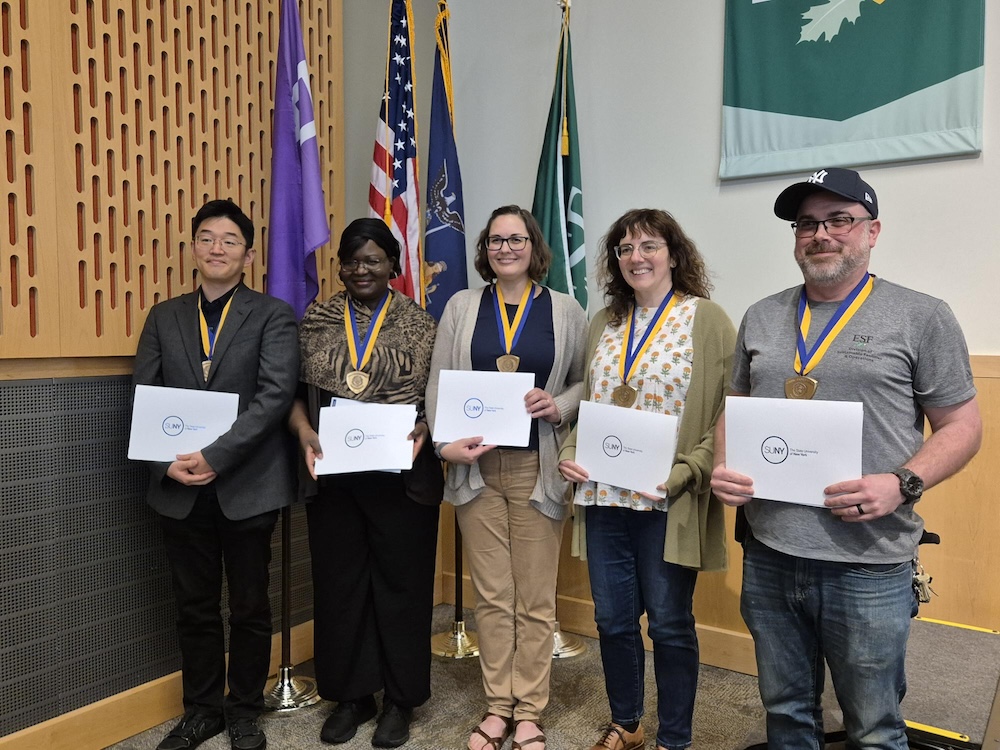 Five people standing in front of flags while holding certificates.