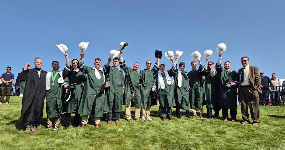 graduates in green robes holding up white construction hats