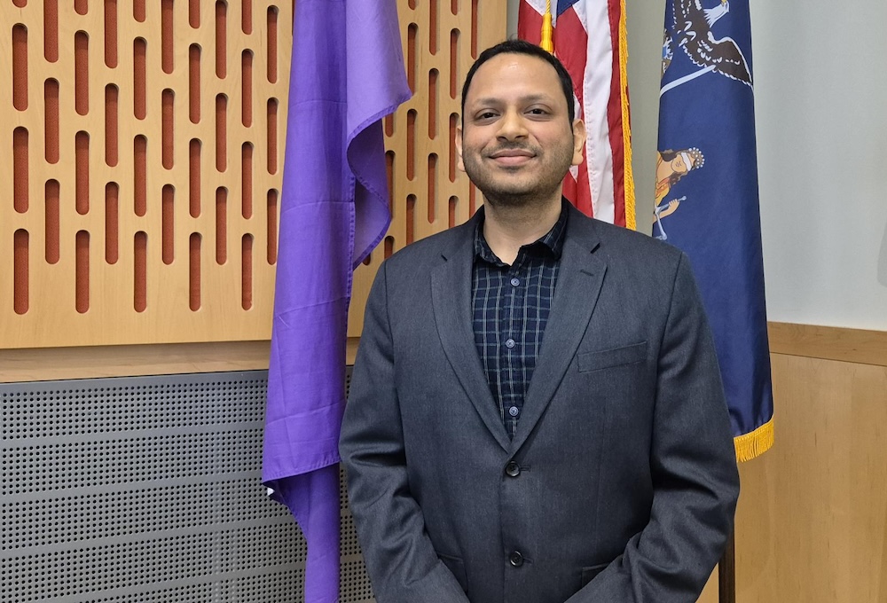 Man in blue suit jacket and blue shirt standing in front of three flags.