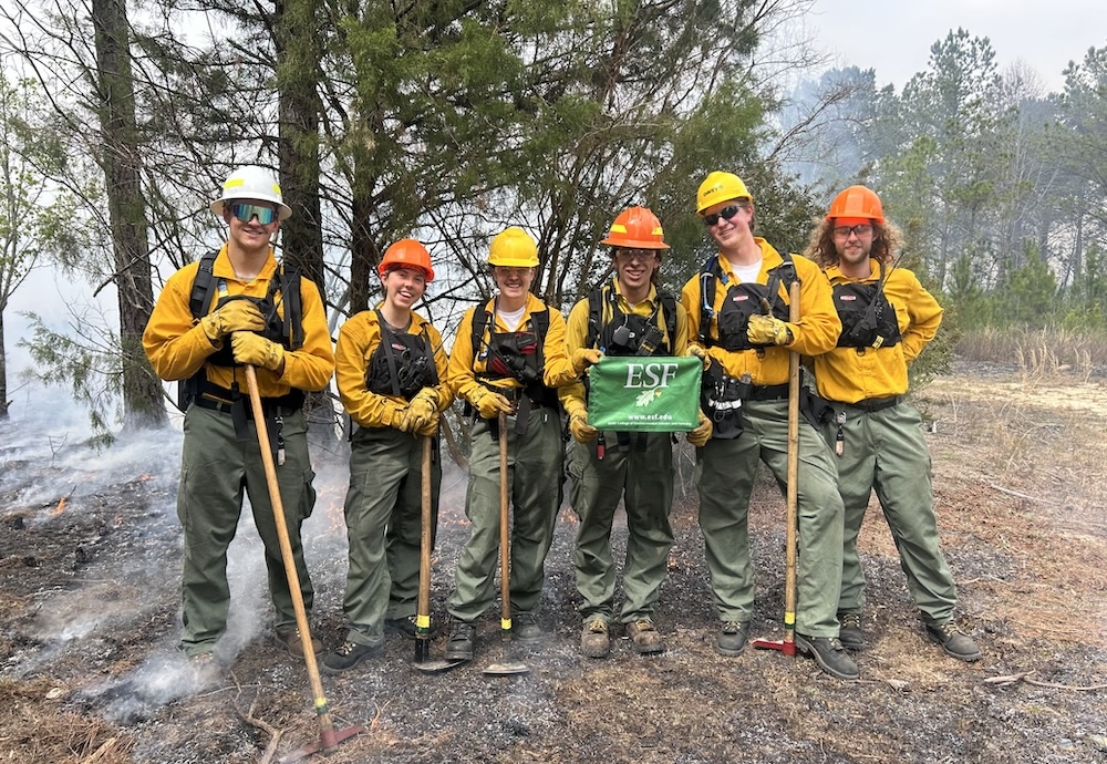 Six people wearing yellow coats, hard hats, and holding axes stand near smokey woods.