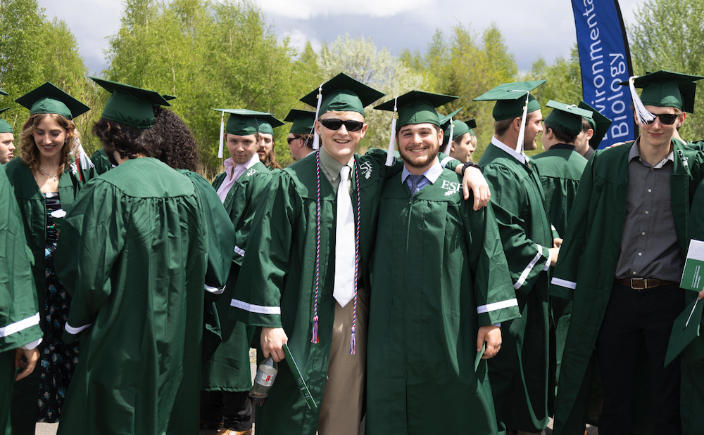 Two men in green regalia with their arms around each other surrounded by other graduates.
