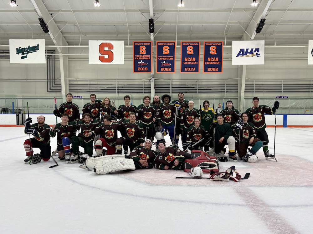 A group of ice hockey players pose together on an indoor rink, wearing matching dark jerseys with a colorful logo, with some kneeling in front and others standing behind. Overhead, banners and logos hang from the ceiling, including a large orange “S” and multiple championship banners displayed along the wall.