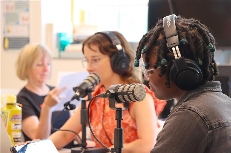 Three people wearing headphones sit at a table with microphones in a recording studio. Two people in the foreground speak into microphones while reading notes, and a third person sits slightly behind them, looking at a sheet of paper. 