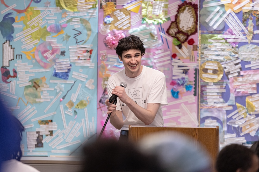 A person stands behind a wooden podium speaking into a handheld microphone. The person wears a light-colored T-shirt and is positioned in front of three large, colorful mixed-media art panels filled with layered shapes, textures, and strips of printed text. Blurred audience members are visible in the foreground, indicating a public presentation or event.