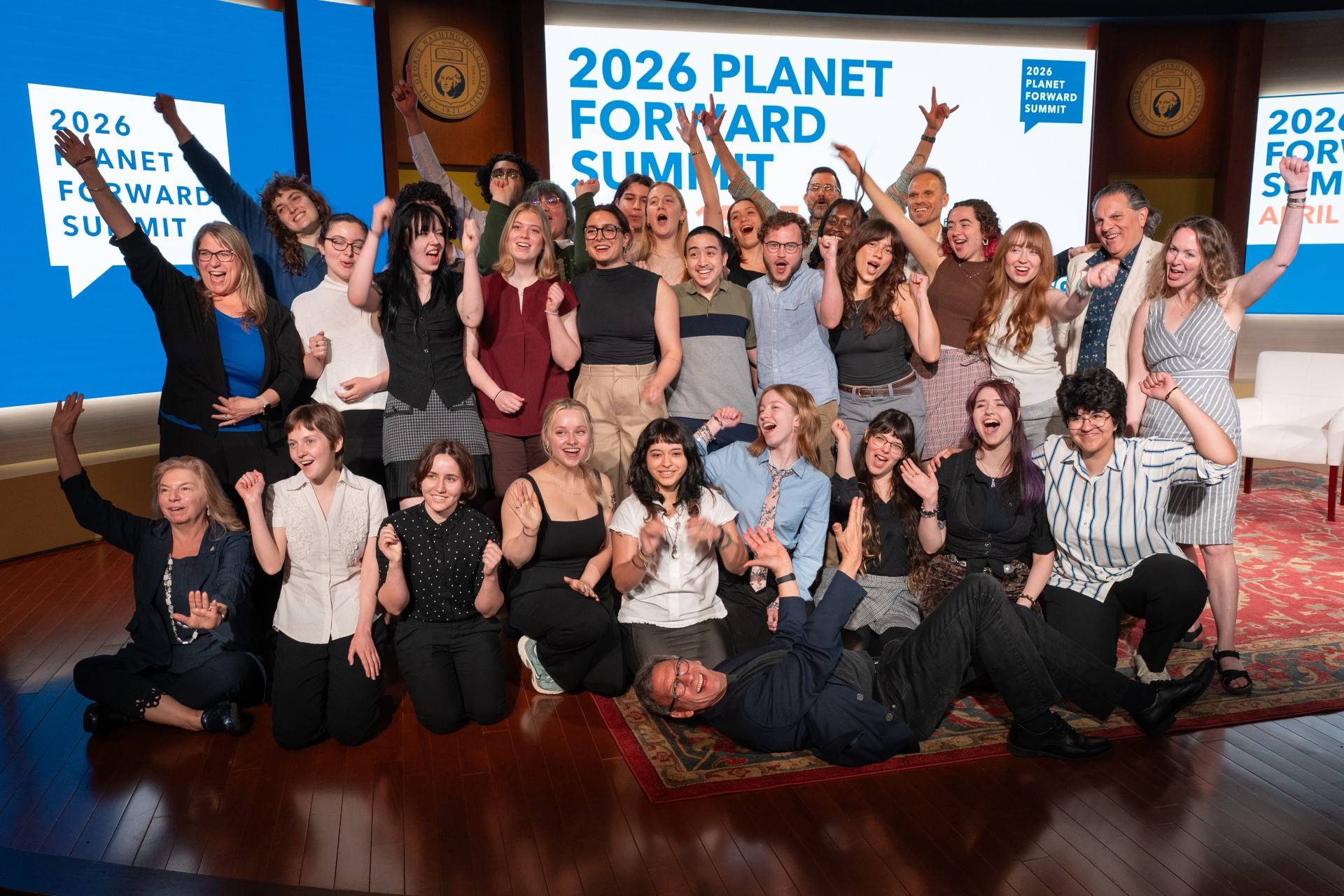 A large group of people pose together on a stage in front of two large screens reading “2026 Planet Forward Summit,” with additional text showing the dates April 15–17. The group is arranged in rows on a patterned rug under stage lighting.