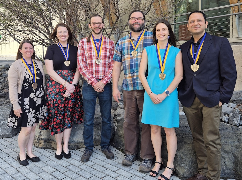 Six people stand outdoors in a row wearing medallions on blue and gold ribbons, posed on a paved walkway in front of a stone wall and a campus building. The group is dressed in professional and business casual clothing, with trees and windows visible in the background.