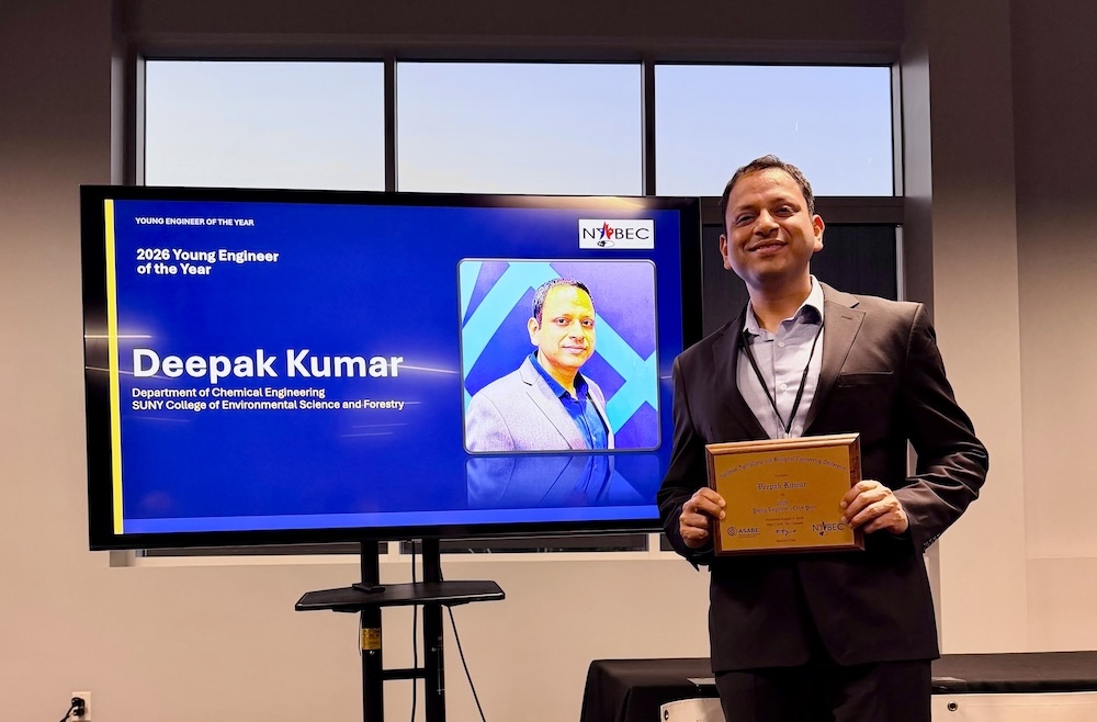 Person holding a plaque stands by a screen with their photo displayed on it and the name Deepak Kumar.