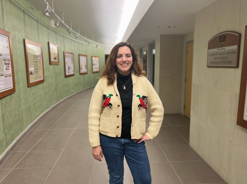 A person stands in a brightly lit hallway lined with framed informational displays on a green textured wall. They are wearing jeans, a dark top, and a cream-colored cardigan with red and green embroidered designs while posing with one hand on their hip.