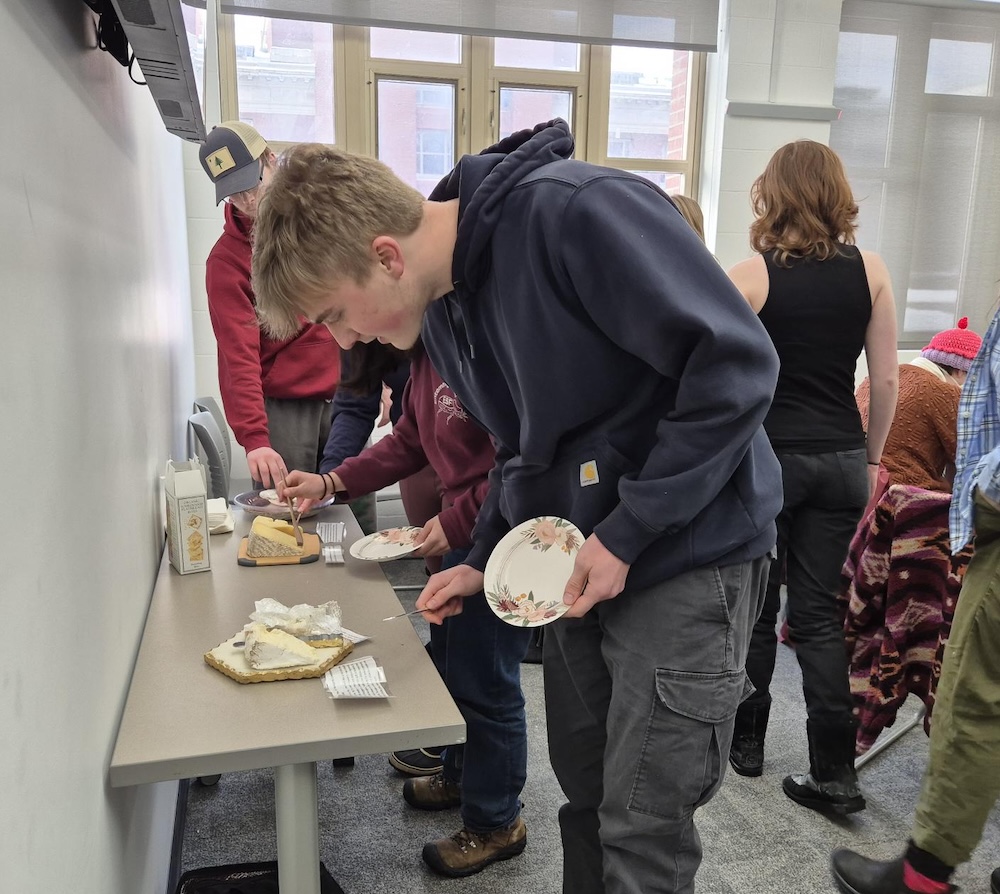 Several people are gathered around a long table in a bright room, serving themselves food. The table holds various trays of cheese and snacks, along with stacks of napkins and small plates. One person in the foreground is leaning forward while holding a decorative paper plate and reaching toward the food. Others stand close by, also selecting items. The room has large windows in the background letting in natural light, and additional people are visible standing and talking near the back of the room.