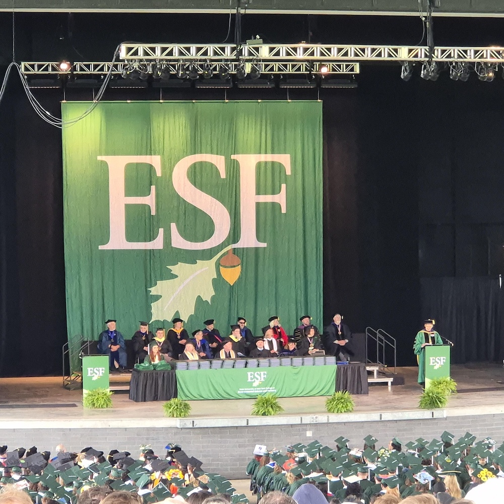 Stage with large ESF banner and people sitting in academic regalia while person in green regalia speaks at podium.