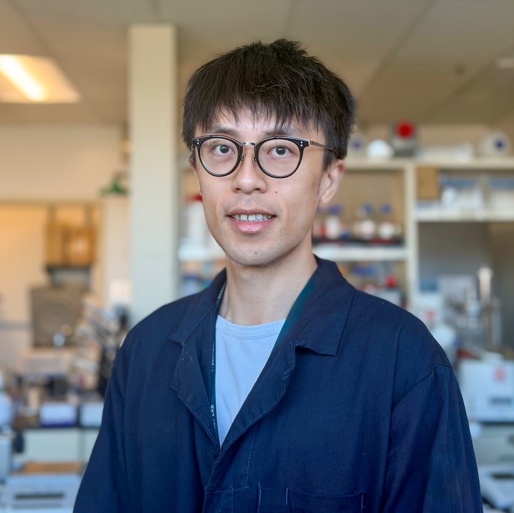 A person wearing a dark lab coat stands in a laboratory setting with shelves of equipment, containers, and instruments visible in the background. The room is brightly lit, showing an organized workspace with scientific supplies arranged along the counters and shelves.