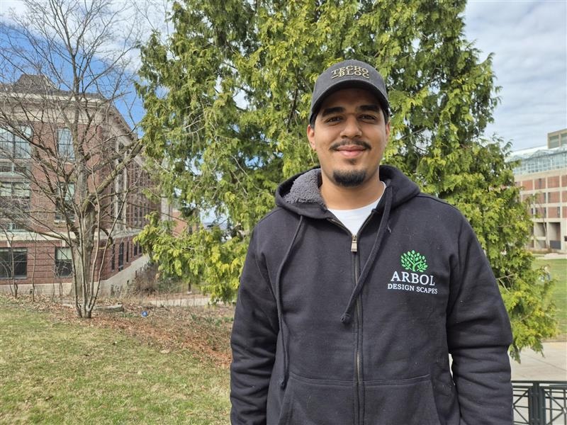 Person in grey hoodie and baseball cap stands in front of a tree.