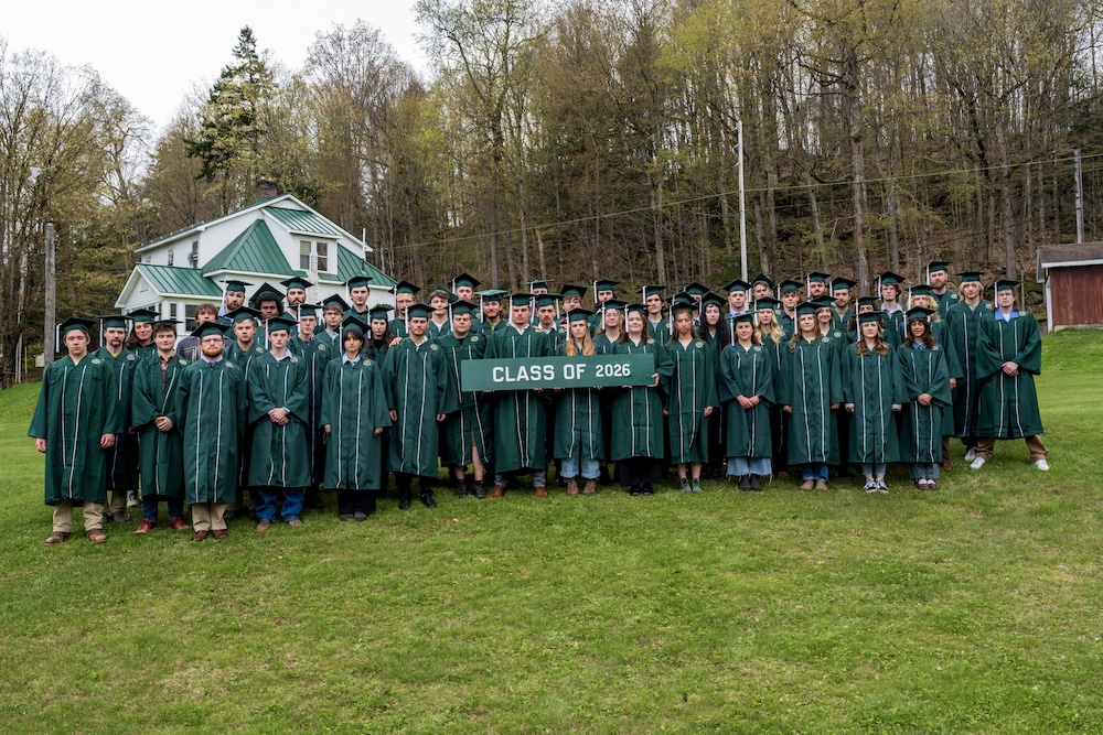 A large group of graduates in green caps and gowns stands on a grassy lawn holding a “Class of 2026” sign. Trees and a small building appear in the background.