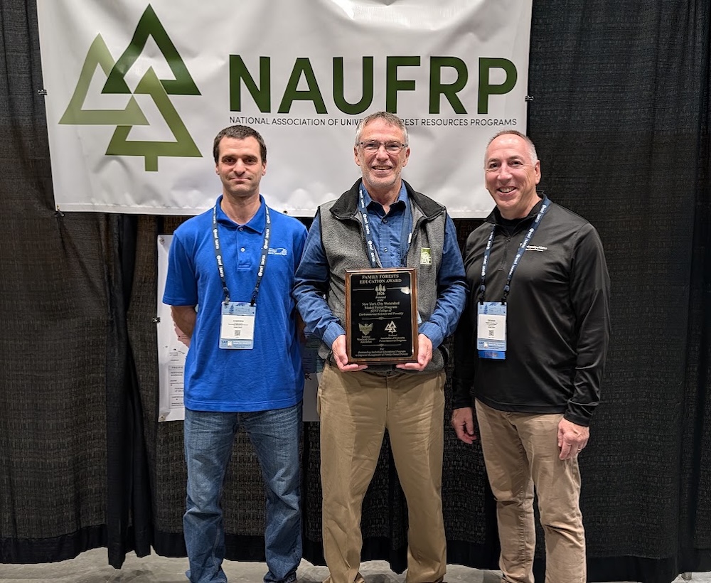 Three people stand in front of a large NAUFRP (National Association of University Forest Resources Programs) banner at an indoor conference or awards event. The person in the center holds a framed plaque recognizing an award, while the two people on either side stand beside the recipient.