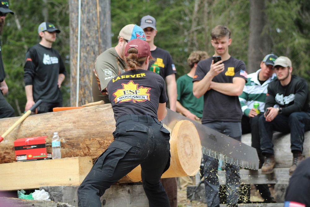 A person uses a large handsaw to cut through a thick log mounted on a stand while wood chips scatter. Several other people stand nearby watching, with trees and outdoor equipment visible in the background.