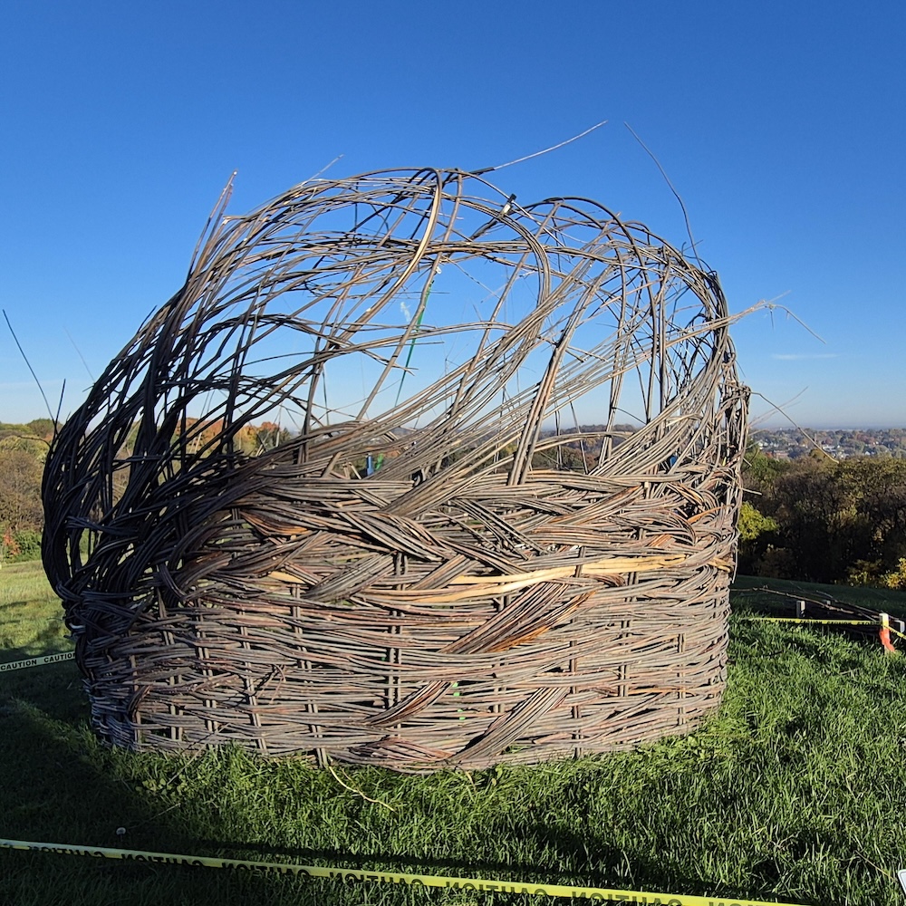 A large, nest-like sculpture woven from long willow branches sits on a grassy hill under a clear blue sky. The structure is cylindrical at the base and rises into an open, swirling form at the top, with branches curving and extending upward in multiple directions. The woven texture creates a basket-like appearance with visible layers of intertwined natural materials.