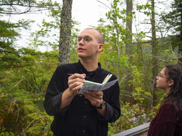 Student looking on as another student reads from a booklet in a wooded setting.