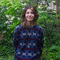 Person in a blue and red patterned sweater with multiple necklaces smiles in front of a background of trees