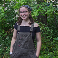 Person with glasses and a blue bandana wearing overalls smiles in front of a background of trees